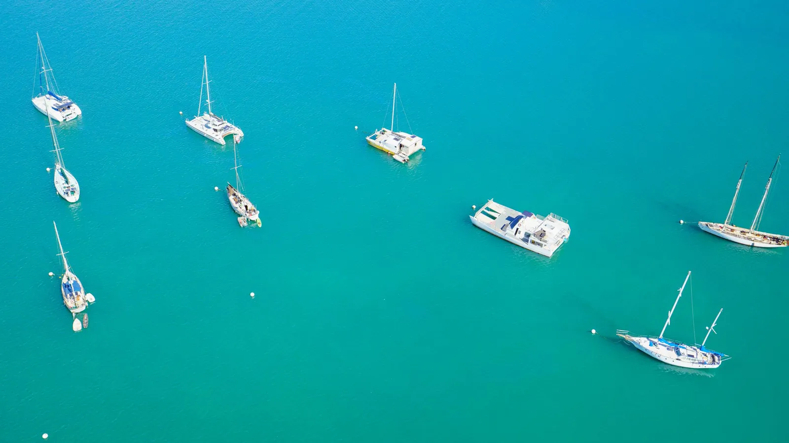 Sailing boats, catamarans and a motorboat lying well apart in a turquoise bay, seen from above.