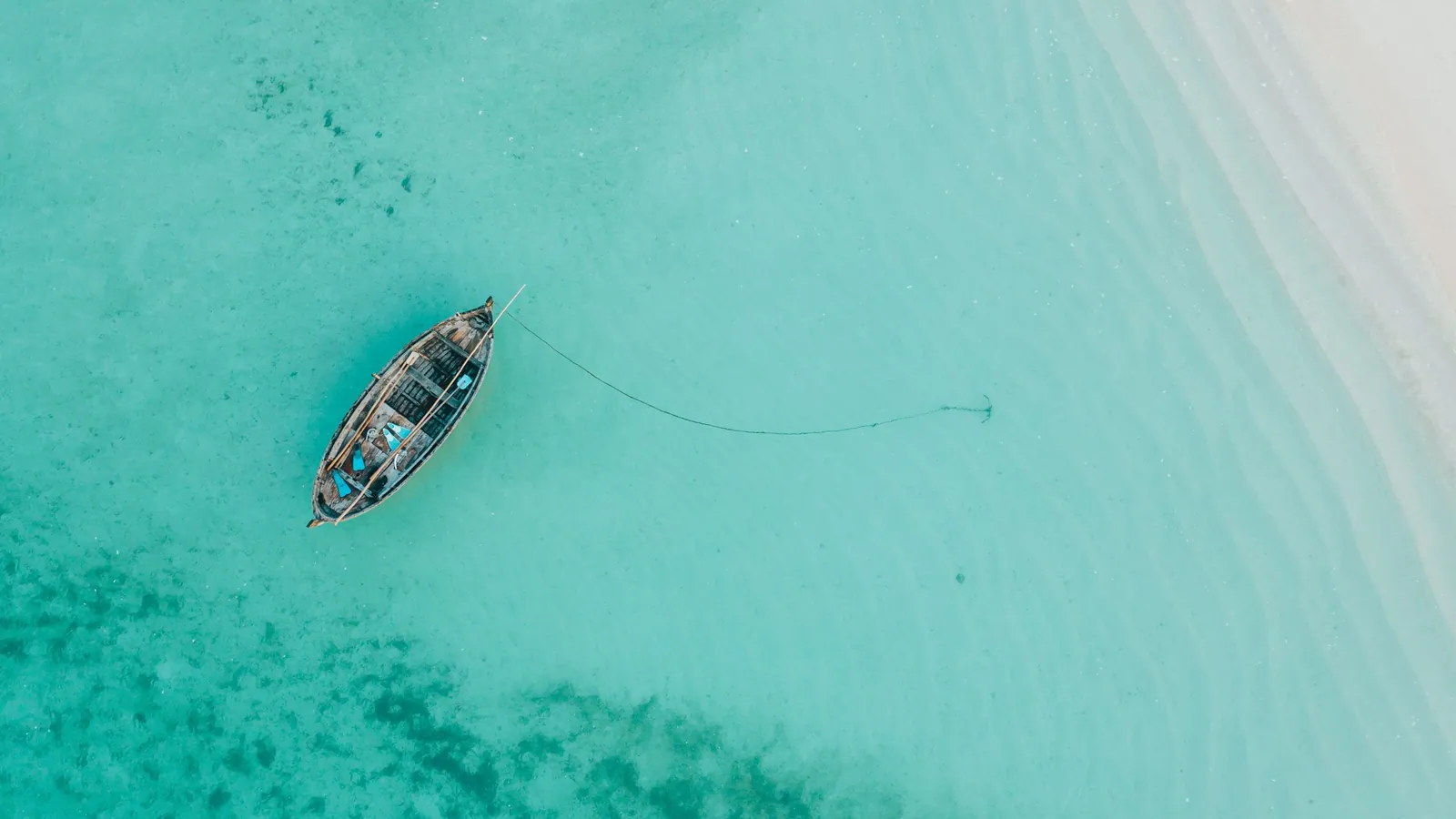 A wooden boat at anchor in clear turquoise water, seen from above. The anchor line runs from the bow across the sandy bottom to the anchor.